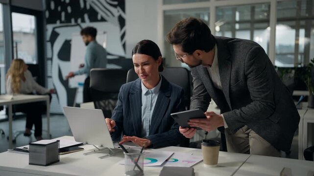 Two Caucasian business people teamwork looking in laptop talking partnership in corporate office European workers partners look in computer and digital tablet talk in company female male cooperate job