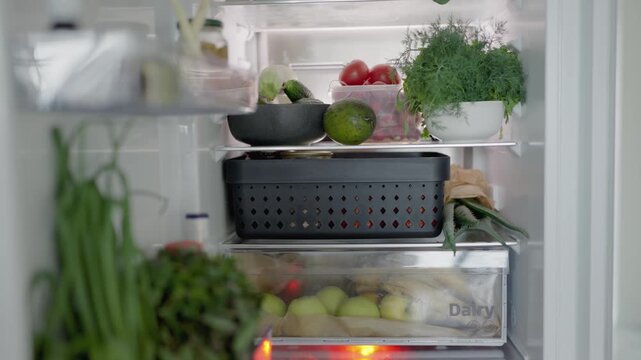 An open refrigerator stocked with a variety of fresh vegetables and fruits, including green herbs, cucumbers, tomatoes, apples, and leafy greens, promoting healthy eating habits.