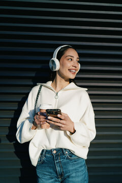 Cheerful young woman with braces using mobile phone in headphones and holding takeaway coffee. Girl enjoying digital connectivity, streaming music or chatting in contemporary urban setting.