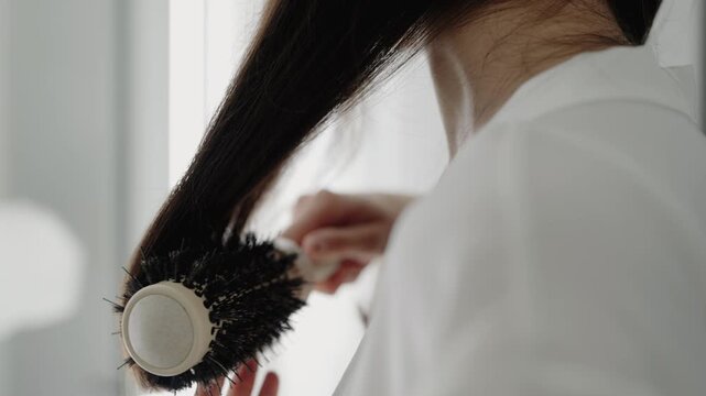 Close-up of a woman brushing her long dark hair with a round brush, wearing a white shirt. Captures daily hair care and grooming routine. Woman Brushing Long Dark Hair With Round Brush in White Shirt