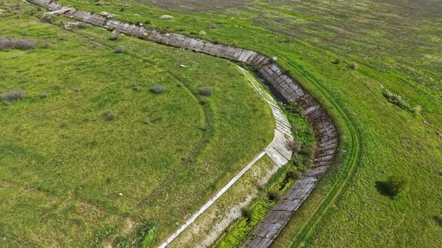Water channels are dry and cracked, showing no water flow. Green grass covers the land around, while gentle hills reshape the rural landscape. Dry soil indicates a lack of moisture.