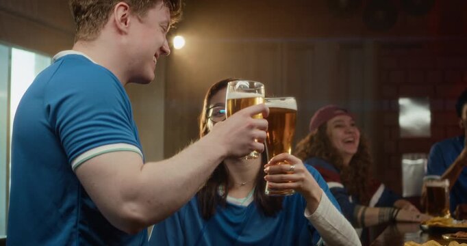 Low angle shot of group of friends in matching shirts clinking beer glasses and watching football game in bar