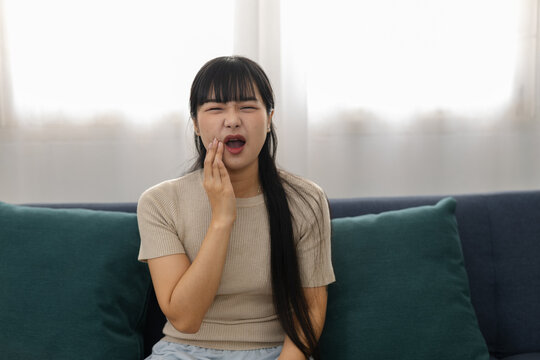 Young adult Asian woman sitting comfortably on modern sofa holding cheek experiencing extreme dental pain showing sad suffering emotion enduring sudden jaw ache inside bright living room
