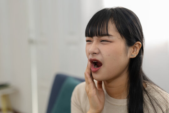 Young asian woman holding jaw with open mouth suffering severe pain and terrible tooth ache needing immediate dental care feeling intense discomfort inside home