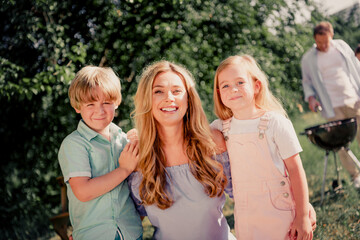 Fototapeta premium Happy family together: mother with son and daughter smiling in sunny backyard while father grills at a summer barbecue