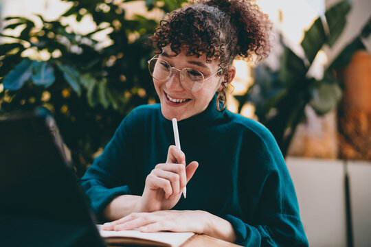 Freelancer woman smiling with pen in hand while brainstorming at tablet and notebook, portraying inspiration and connection between ideas and tech tools in a flexible digital workflow.