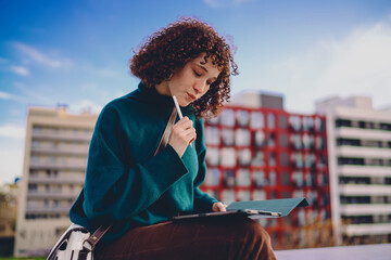 Low angle view of young woman holding stylus to lips, deeply focused on digital tablet, highlighting modern tech thinking, urban creativity and mobile freelance lifestyle. © BullRun
