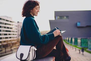 Side view of smiling woman drawing on tablet, relaxed posture showing comfort and focus of freelance tech lifestyle where creativity flows freely in urban open-air environment. © BullRun