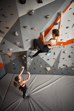 Woman supports climbing partner during bouldering session indoors. Photo for safety, encouragement, teamwork, strength, gym membership marketing, sport communication