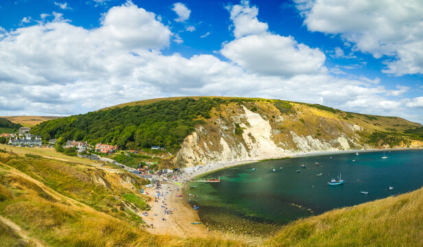 An expansive summer landscape of a natural cove showing holiday accommodations, a crowded beach, and white chalk cliffs meeting the ocean water (Lulworth, England, United Kingdom)