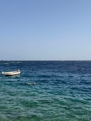 Obraz premium A small white boat floats on clear blue water under a bright sky. The scene captures a serene coastal landscape with gentle waves.
