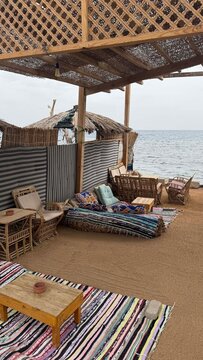A beachside lounge area with wooden furniture, colorful rugs, and a view of the sea. The setting is relaxed and inviting, perfect for leisure and relaxation.