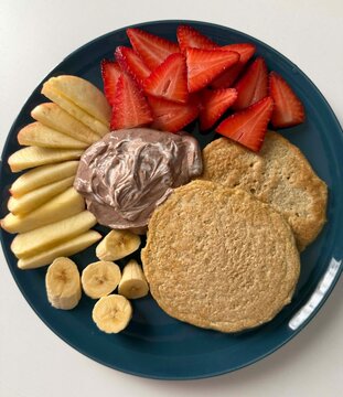 A blue plate filled with healthy breakfast items. Includes sliced apples, strawberries, banana slices, and two round pancakes with chocolate spread.