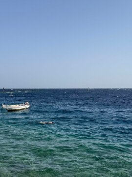 A small white boat floats on clear blue water under a bright sky. The scene captures a serene coastal landscape with gentle waves.