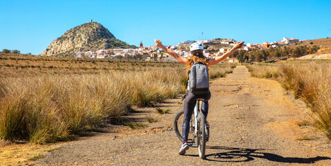 Fototapeta premium happy young woman cyclist with backpack raising arms in joy while riding a bicycle on a rural road in spain