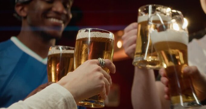 Low angle close up shot of group of friends clinking glasses of draft beer, while watching sports game at bar