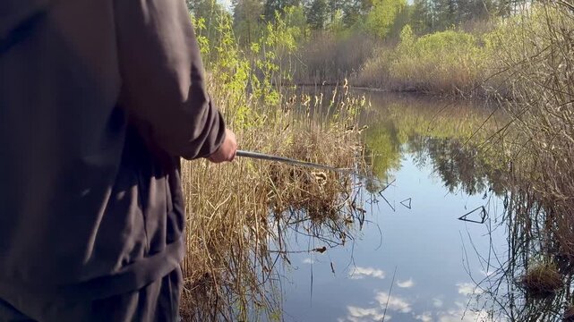 A fisherman standing on the shore with a fishing rod, watching the float on the water. The video is filmed from behind. The fisherman's face is not visible. The water is calm