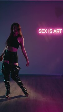 A brunette woman dances in black high boots and shorts. Neon light on wall display. Gray background. Purple light illuminates scene. Sensual Dance Performance