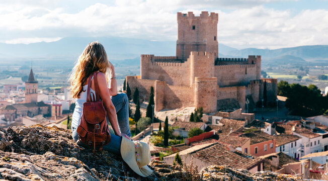 young woman with backpack sitting on a hill and contemplating the panoramic view of the medieval atalaya castle in villena, spain