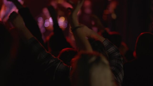 Fans raise hands and smartphones in concert crowd, teal blue stage light illuminates crowd from front, phones visible throughout scene