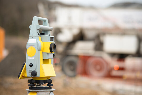 Survey equipment stands in focus at a construction site with machinery in the background on a cloudy day