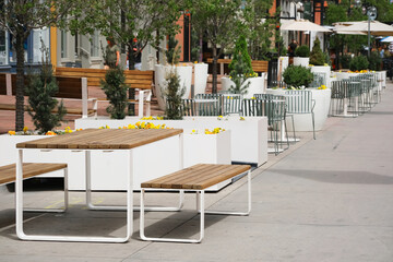 Outdoor cafe with white planters, wooden benches and metal tables lining a sunlit urban pedestrian plaza and wide sidewalk, contemporary terrace seating. © elena.tres