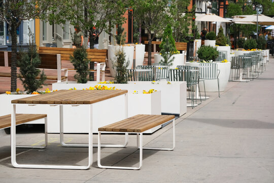 Outdoor cafe with white planters, wooden benches and metal tables lining a sunlit urban pedestrian plaza and wide sidewalk, contemporary terrace seating.