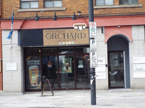 Gatineau, Quebec, Canada &ndash; March 10, 2026: "Orchard Pub & Grill" features a brick facade, large windows with beer posters, a blue flag, and several street parking signs at 131 Promenade du Portage.