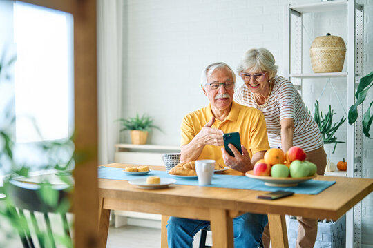 Portrait of an elderly senior couple having breakfast and using a smart phone at home. Happy healthy affectionate senior couple talking and showing smartphone screen to one another and texting and sit