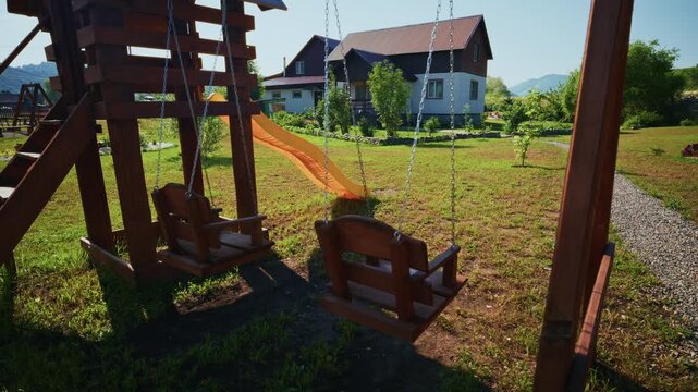 Wooden swing set stands in a grassy yard. Swings hang still under bright sunlight. House with a sloped roof appears in background. Gravel path leads through green lawn. Scene feels calm and peaceful