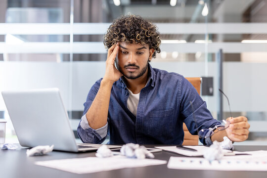 Thoughtful young Indian man sitting at a desk in the office, holding his head in his hands and looking at the crumpled paper in frustration