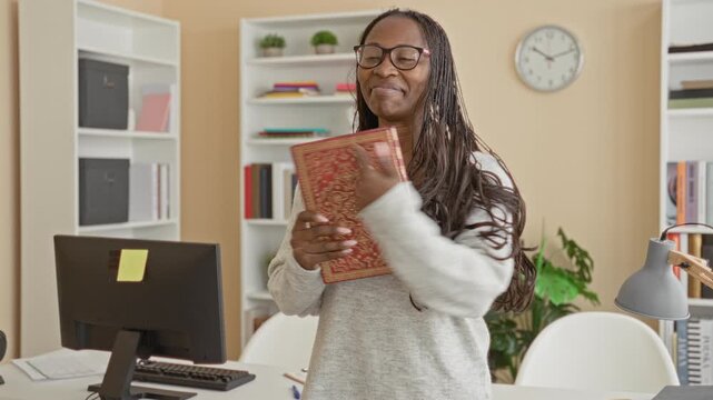 Woman hugging ornate book with hands over chest, smiling and wearing glasses and sweater at a tidy office desk with computer and bookshelves; contentment.