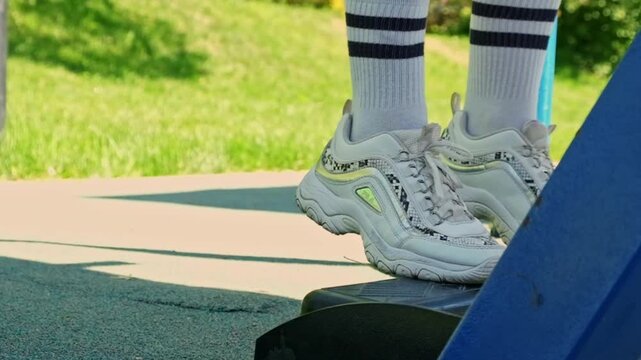 A low close-up shows sneakered feet lightly shifting on playground equipment, with bright grass behind and a relaxed summer rhythm. Use it for outdoor play, active lifestyle, childhood, and wellness.