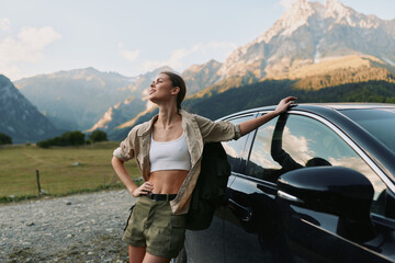 Fototapeta premium Young woman in casual outfit leans on car outdoors enjoys fresh air with mountains in background expresses freedom and lifestyle in nature travel concept.