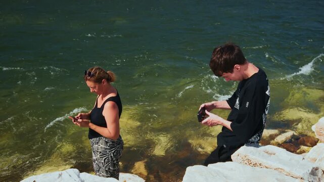 Two people examine small objects near water's edge. They focus intently on their finds. Sunlight glints off gentle waves. Rocks line the shoreline. Ideal for travel, nature, or exploration themes