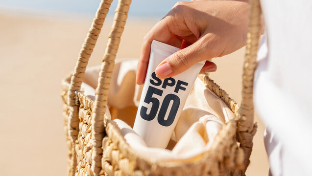 Close-up of hand placing SPF 50 sunscreen tube into woven beach bag, warm golden sunlight, sandy background and natural tones highlighting summer skincare, sun protection and travel preparation