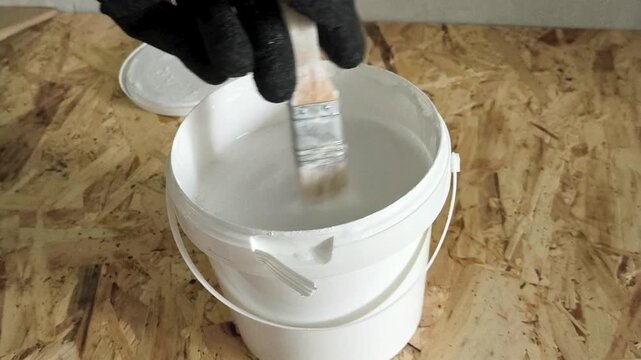 A painter's hand in a glove uses a brush to stir the white paint in the jar.
