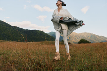 Woman in a light sweater and coat stands in a grassy field near mountains, enjoying fresh air and outdoor lifestyle amid nature. © SHOTPRIME STUDIO