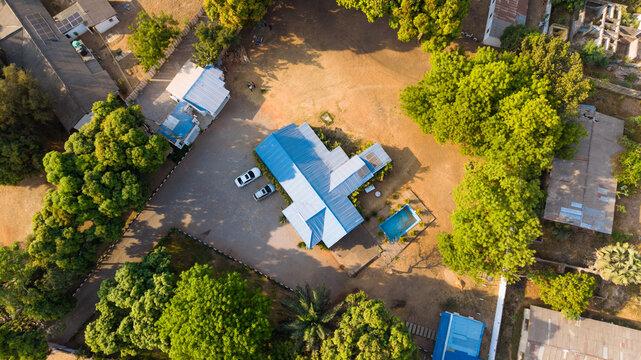Aerial view of a modern building with a blue roof, parked cars, and a swimming pool surrounded by lush green trees and sandy terrain in Kaduna, Kaduna, Nigeria.