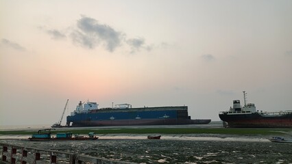 Large cargo ships anchored offshore at sunset with debris in the foreground, industrial seascape © Abdur