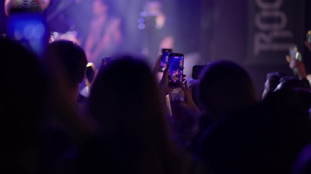 Fan in concert crowd holds smartphone above head to film, phone screen visible, teal blue atmosphere fills venue, performer visible behind