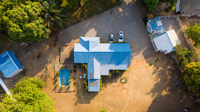 Aerial view of a residential building with a blue corrugated metal roof, a small swimming pool, and parked cars surrounded by lush green trees in Kaduna, Kaduna, Nigeria.