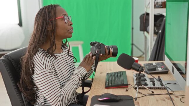 Woman holding camera and gesturing with hand at desk in studio with green screen, microphone and computer visible; focus creativity.
