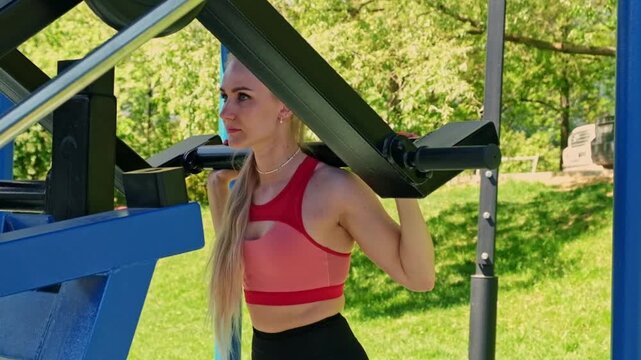 A focused young woman performs a squat on outdoor gym equipment, framed by blue metal bars and green trees in bright light. It suits fitness, strength training, wellness, discipline, and active
