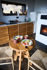 Naklejka premium Healthy breakfast setup in cozy modern kitchen with fireplace, wooden table set for two with eggs, avocado, prosciutto and coffee, concept of home lifestyle, comfort, slow living