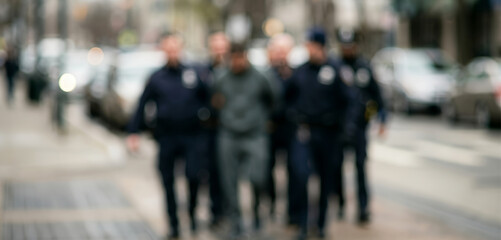 Obraz premium Blurred urban setting depicts law enforcement escorting a suspect on a sidewalk, with indistinct figures and vehicles reinforcing privacy, tension, and everyday policing activity.