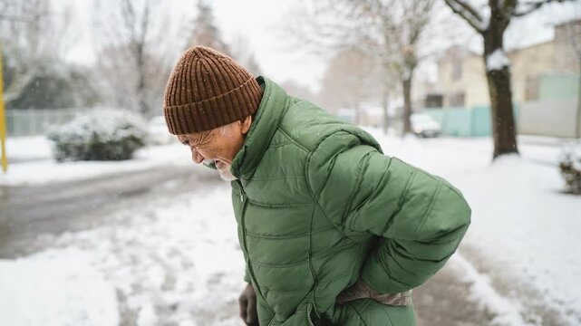Elderly man in green jacket and brown hat suffering back pain outdoors in snowy winter street