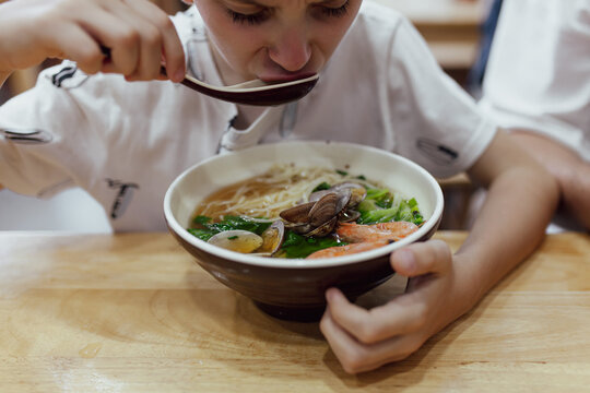 Close-up of boy eating seafood noodle soup ramen with clams, shrimp and fresh greens from bowl. Asian seafood cuisine for children, comfort dining, fresh ingredients.