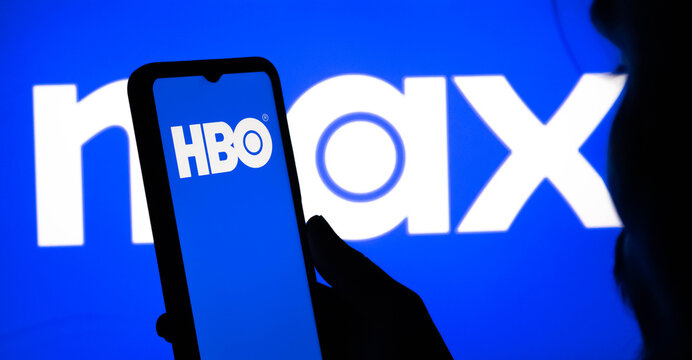 Person holding a smartphone displaying the hbo logo in front of a large max streaming service logo on a screen