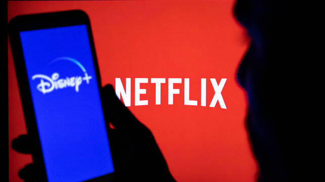 Closeup of a person holding a smartphone displaying the disney plus logo with the netflix logo in the background on a screen